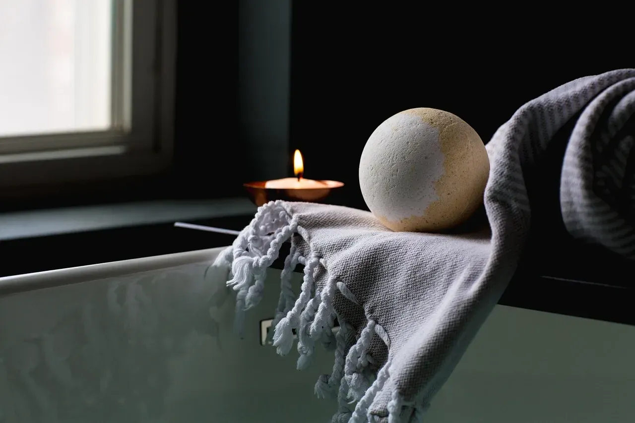 A towel, a bath bomb and a lit candle on the edge of a bathtub.