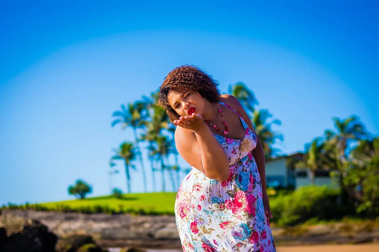 A woman in a floral summer dress blowing a kiss in front of palm trees.